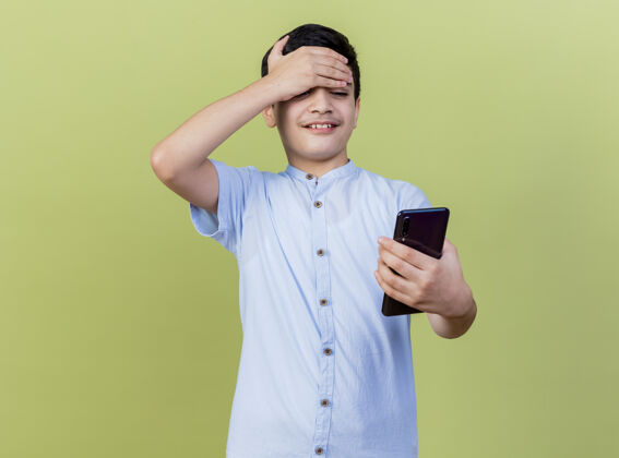 抱着微笑着的年轻白人男孩拿着手机 看着手机 手放在额头上 在橄榄绿的背景上 留着空白手机微笑年轻