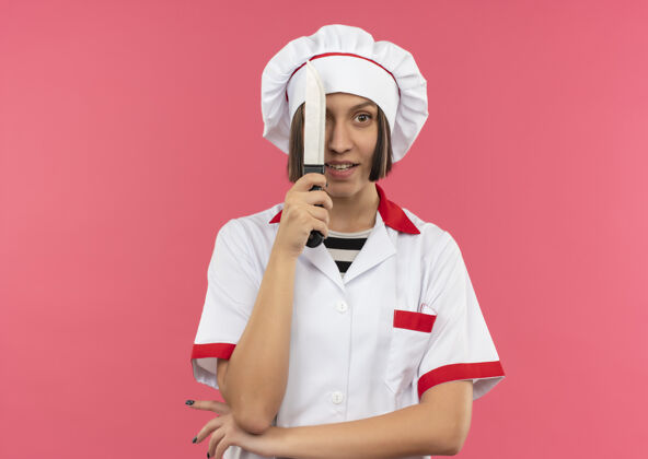 印象令人印象深刻的年轻女厨师在厨师制服从刀后面看 把手肘下孤立的粉红色与复制空间手女空间