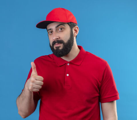 自信自信的年轻英俊的白人送货员穿着红色制服 戴着帽子 在蓝色的地板上竖起大拇指帽子穿英俊
