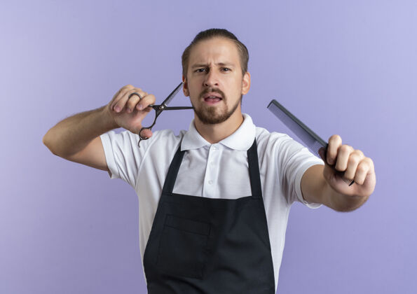 制服印象深刻的年轻英俊的理发师穿着制服伸出梳子和剪刀孤立的紫色伸展紫色剪刀