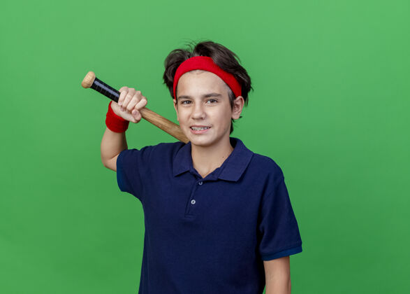 持有面带微笑的年轻帅气的运动男孩戴着头带和戴着牙套的腕带看着相机拿着棒球棒在绿色背景上与复制空间隔离帅气年轻运动