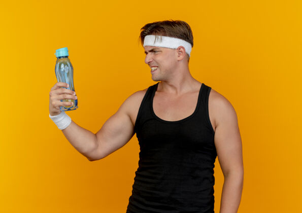 抱着恼怒的年轻英俊的运动男子戴着头带和腕带拿着和看着孤立在橙色的水瓶水男人头带