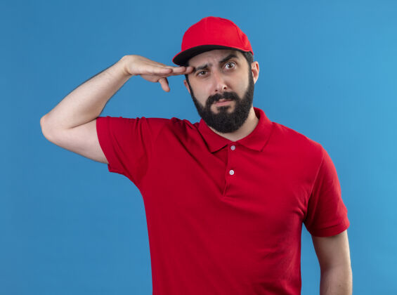 制服自信的年轻英俊的白人送货员身穿红色制服 戴着帽子 手靠近脑袋 孤立在蓝色上近头男人