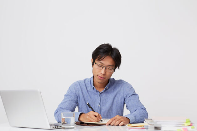 笔记本电脑体贴专注的亚洲年轻商人戴着眼镜 拿着笔记本在桌边工作 在白墙上的笔记本上写计划专业年轻自信
