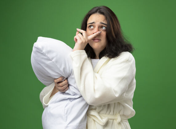 湿巾虚弱的年轻白种人生病的女孩穿着长袍站在侧视图拥抱枕头看着一边擦鼻子隔离在绿色背景与复制空间壁板站着长袍