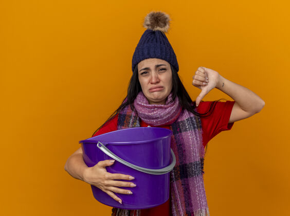 不愉快年轻的高加索病女孩戴着冬天的帽子和围巾拿着塑料桶恶心看着相机显示拇指向下孤立在橙色背景与复制空间女孩向下围巾