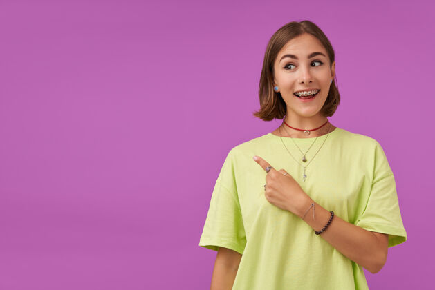 项链十几岁的女孩 快乐快乐 深色短发 手指指向紫色墙上的复印处展示一个标志穿着绿色t恤 手镯和戒指年轻牙套微笑