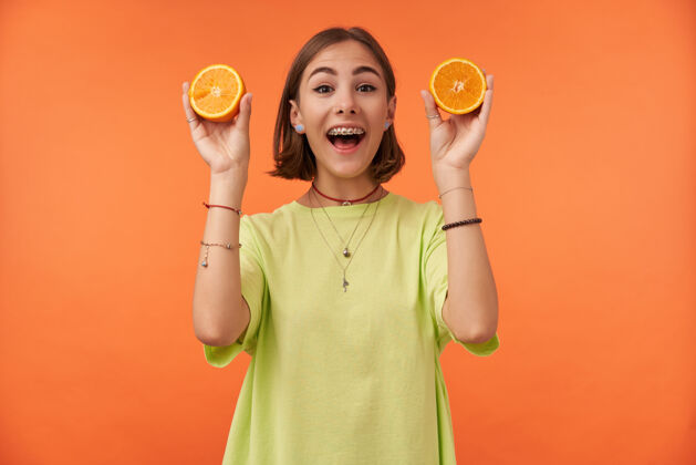牙齿漂亮漂亮的女孩的肖像 深色短发 橘子色 微笑准备做果汁穿着绿色t恤 牙套和手镯年轻人女孩时髦
