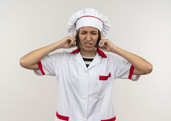 耳朵恼怒的年轻女厨师穿着厨师制服把手指放在耳朵上闭着眼睛孤立在白色女性手指烹饪