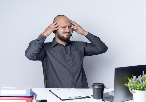 工作疼痛的年轻秃头呼叫中心男子戴着耳机坐在办公桌前 一边看着工作工具 一边把手放在头上 隔离在白色的坐着穿手