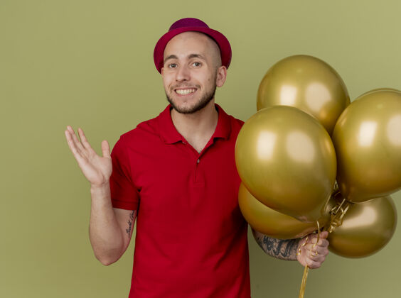 手面带微笑的年轻英俊的斯拉夫党人戴着党的帽子拿着气球看着相机显示空手孤立在橄榄绿的背景帅气年轻相机