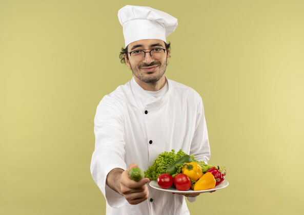 男年轻的男厨师穿着厨师制服 戴着眼镜 手里拿着盘子里的蔬菜 手里拿着隔离在绿墙上的黄瓜黄瓜盘子制服