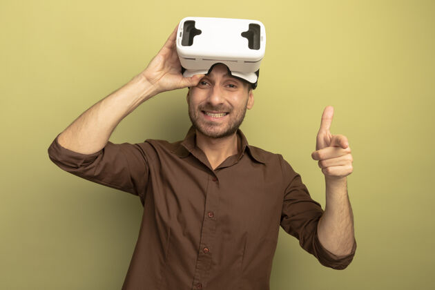 橄榄微笑的白人年轻人头戴虚拟现实耳机抓住它看着并指着橄榄绿背景上孤立的摄像头头虚拟现实年轻