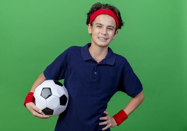 帅气面带微笑的年轻帅气的运动男孩戴着头带和戴着牙套的腕带拿着足球手放在腰上看着隔离在绿色背景和复制空间的相机手举行背带
