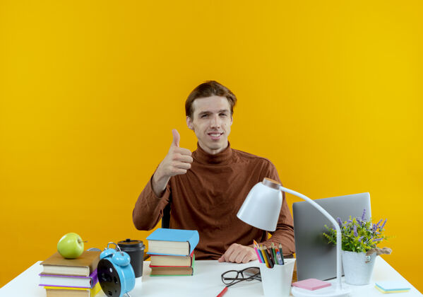 学生高兴的年轻学生男孩坐在书桌旁 手里拿着学习工具 他的大拇指孤立地竖在黄色的墙上工具学校男孩
