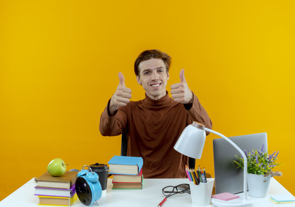 坐着微笑的年轻学生男孩坐在书桌旁 手里拿着学习工具 他的大拇指孤立地竖起在黄色的墙上工具拇指男孩