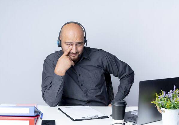 电话自信的年轻秃头呼叫中心男子戴着耳机坐在办公桌旁 用工作工具把手指放在眼睛下面 隔离在白色上坐着穿中心
