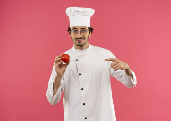 请高兴的年轻男厨师穿着厨师制服 拿着眼镜 指着隔离在粉红色墙上的西红柿眼镜男烹饪