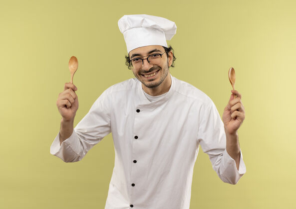 年轻面带微笑的年轻男厨师穿着厨师制服 戴着眼镜 拿着勺子孤立地站在绿色的墙上厨师微笑勺子