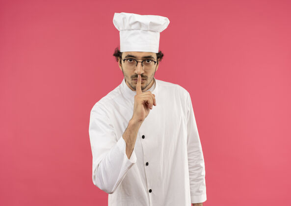 穿着穿着厨师制服 戴着眼镜的严格的年轻男厨师在粉红色的墙上表现出沉默的姿态烹饪制服表演