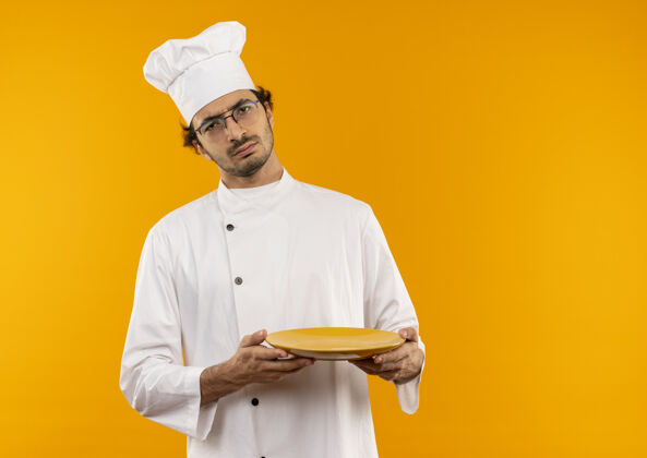 盘子严格年轻的男厨师穿着厨师制服 戴着眼镜把盘子隔离在黄色的墙上严格男性年轻