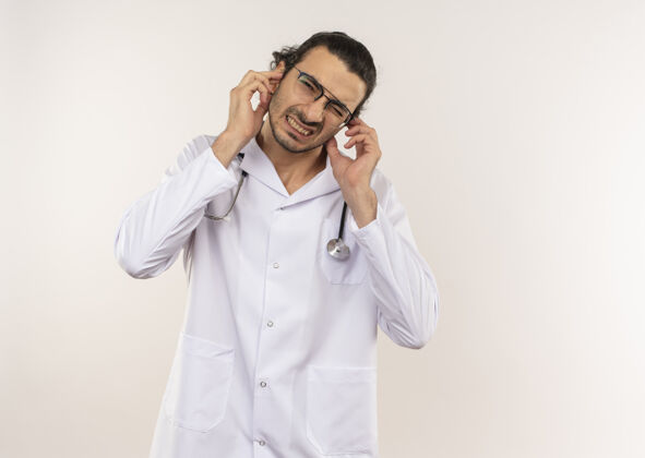 封闭神经质的年轻男医生 戴着眼镜 穿着白色长袍 带听诊器 在隔离的白色墙壁上闭着耳朵 留着复印空间眼镜戴医生