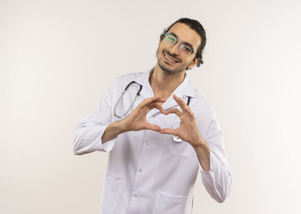 心脏微笑的年轻男医生 戴着眼镜 穿着白色长袍 听诊器 在隔离的白色墙壁上显示着心脏的姿势 还有复印空间手势微笑表演
