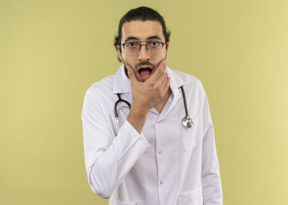 听诊器惊讶的年轻男医生戴着眼镜 穿着白色长袍 听诊器 手放在下巴上 隔离的绿色墙壁上 留有复印空间光学男性年轻