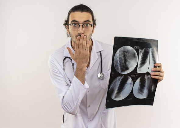 戴年轻的男医生戴着眼镜 身穿白色长袍 听诊器拿着x光片 手捂着嘴 站在隔离的白墙上 留着复印空间罩子听诊器X光