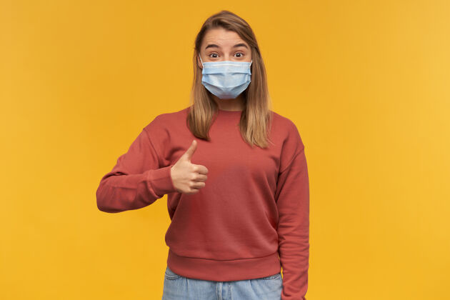 医学快乐兴奋的年轻女子 戴着防病毒口罩 站在黄色的墙上 竖起大拇指快乐流行病女人