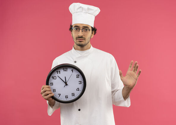 制服悲伤的年轻男厨师穿着厨师制服 戴着眼镜 手里拿着挂钟 把手孤立地摊开在粉红色的墙上厨师持有钟