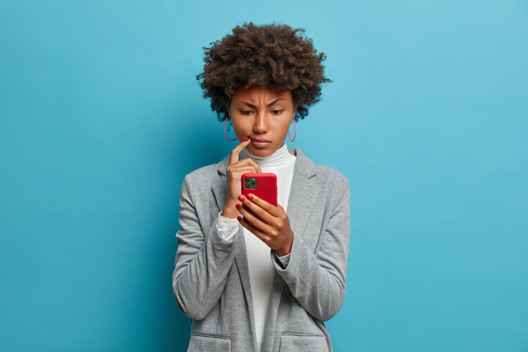 民族迷惑不解的黑皮肤正装女子怒视着智能手机 凝视着显示器 在网站上阅读商业新闻 应用程序女人女士