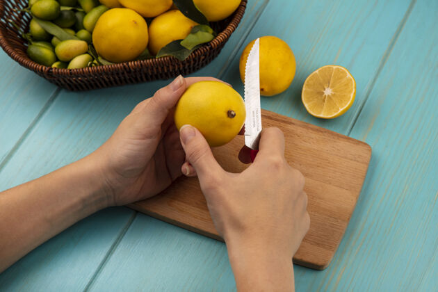 木头女性用刀在木制厨房板上切黄皮柠檬的手俯视图 蓝色木制墙上的桶上放着金盏花和柠檬等水果皮肤顶部视图