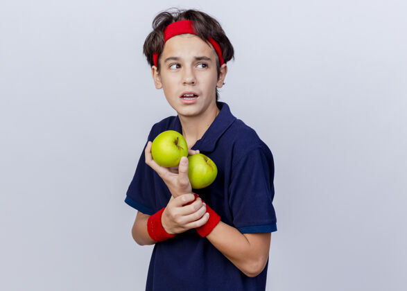 苹果令人印象深刻的年轻英俊的运动男孩戴着头带和戴着牙套的腕带站在侧视图中拿着苹果和手腕看着白色背景上与副本空间隔离的一面白色腕带持有