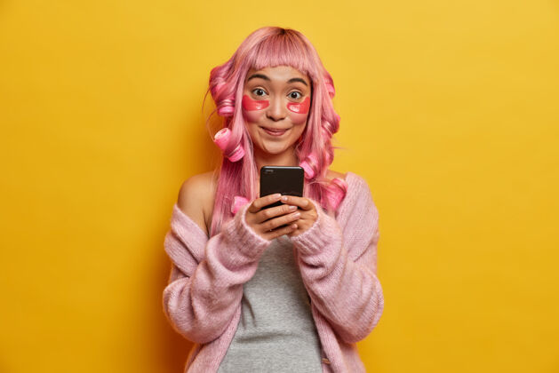 完美微笑积极的亚洲女人手里拿着手机 查看邮箱 神采奕奕 有着粉红色的头发 戴着滚轴检查纯洁黄色