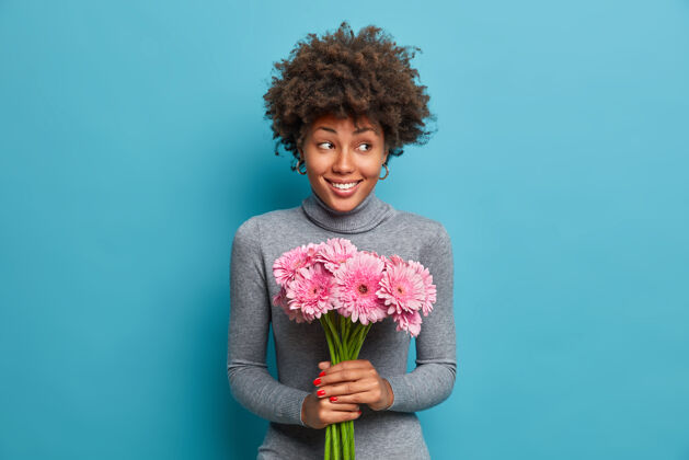 快乐开朗的黑皮肤女士手持粉色非洲菊的花束 把喜悦和幸福放在一边 穿着灰色高领毛衣满意室内请