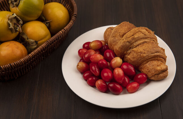 风景白色盘子上的羊角面包配上红酸的山茱萸樱桃 木桶上的柿子水果的俯视图桶酸的水果