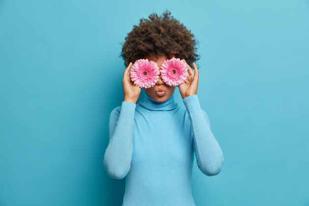 时尚有趣的卷发年轻女子用粉色非洲菊雏菊遮住眼睛 为朋友做花束和最好的自然礼物 穿蓝色高领毛衣花店生涯美丽的花朵 宜人的香味开花女性季节