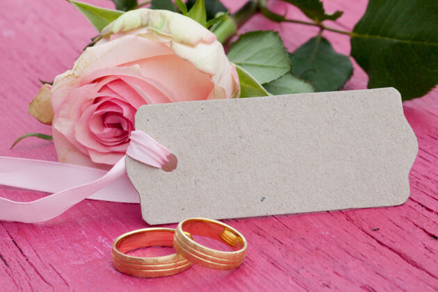 戒指一支粉色玫瑰的特写镜头 一个有文字空间的标签和一张粉色桌子上的两枚金色结婚戒指情侣心宏