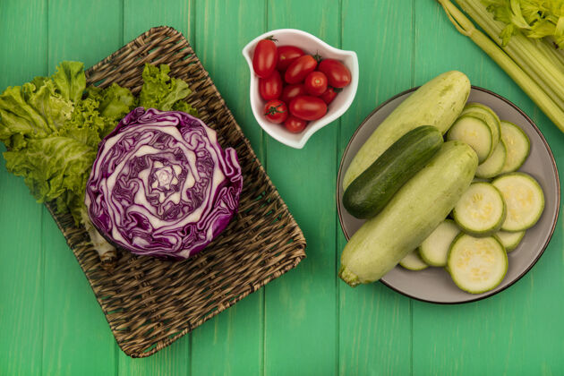 李子紫色卷心菜放在柳条托盘上 莴苣 西葫芦和黄瓜放在盘子上 西红柿放在碗里 芹菜被隔离在绿色的木墙上黄瓜卷心菜柳条