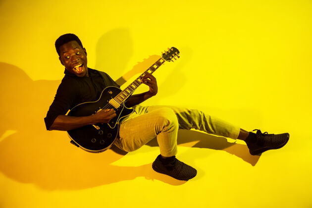 白天年轻的非洲裔美国音乐家在霓虹灯下像摇滚明星一样在黄色背景下弹吉他艺术家俱乐部吉他手