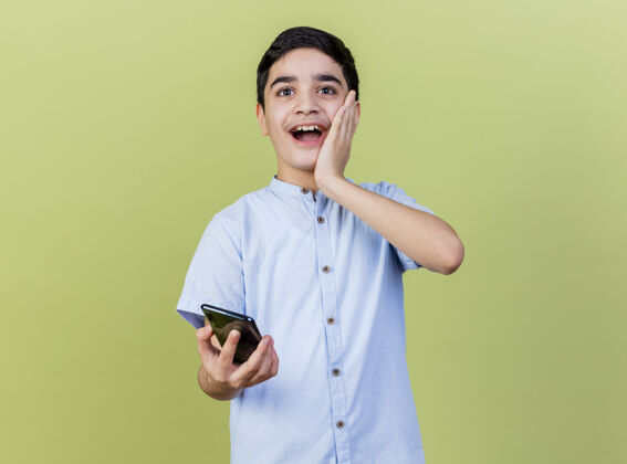 年轻人惊讶的小男孩拿着手机看着前面 手放在脸上 隔离在橄榄绿的墙上市民手机感觉