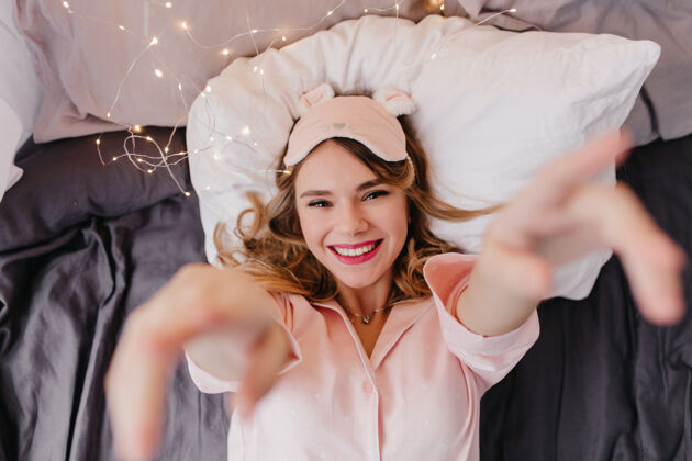 唤醒快乐的金发女孩躺在深色床单上室内头顶照片微笑的快乐女士戴着粉红色的眼罩在床上摆姿势年轻成人女人