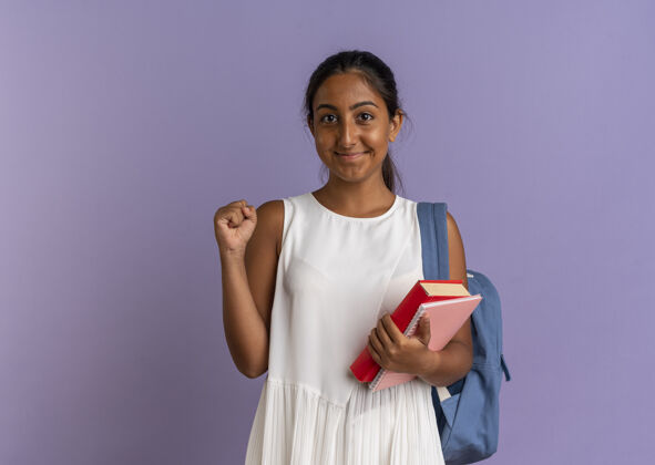 笔记本高兴的小女生背着书包拿着书和笔记本 在紫色上显示“是”的手势拿着请紫色