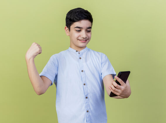 握着微笑的小男孩抱着手机 看着橄榄绿墙上做着强烈的手势电话脸姿势