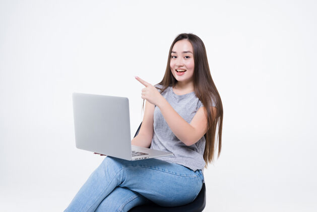 美丽一个快乐随意的亚洲女人坐在白色墙壁上的椅子上 一边拿着笔记本电脑一边指着画像快乐自信椅子