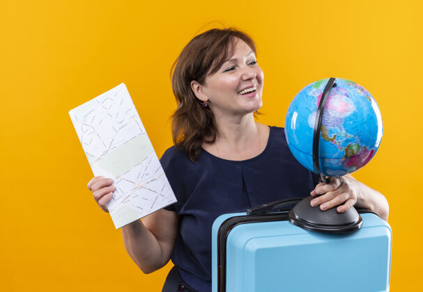 地球仪看着一旁微笑着的中年旅行家妇女 手里拿着手提箱和地球仪 孤零零的黄色墙上挂着地图壁板手提箱中年人