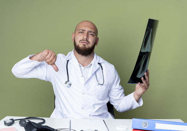 按住年轻的秃头男医生穿着医用长袍和听诊器坐在办公桌上工作长袍向下医疗