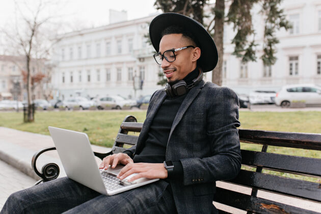 英俊严肃的非洲男人穿着黑色衬衫和裤子在露天使用笔记本电脑户外照片的黑白混血儿自由职业者休息在公园长凳上笔记本电脑年轻学习