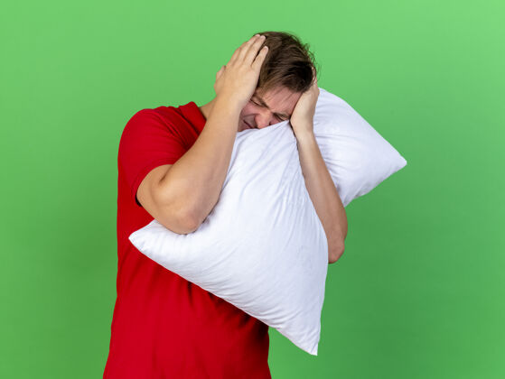 枕头疼痛的年轻英俊金发病男子抱着枕头双手放在头上头痛闭着眼睛隔离在绿色的墙上感觉疼痛抱着